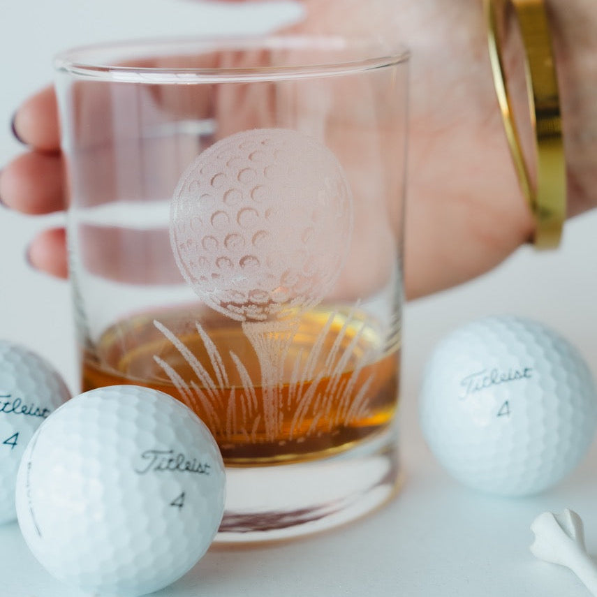 Clear glass mug with golf ball design filled with liquid, surrounded by golf balls on a white surface.