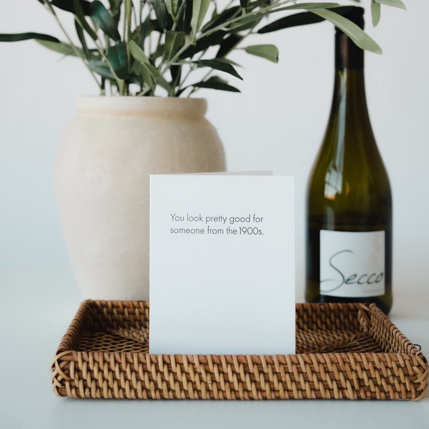 Vase with greenery, bottle of wine, and card on a tray against a light background