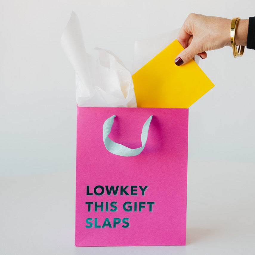Pink gift bag with 'LOWKEY THIS GIFT SLAPS' text, yellow gift card, and white tissue paper.