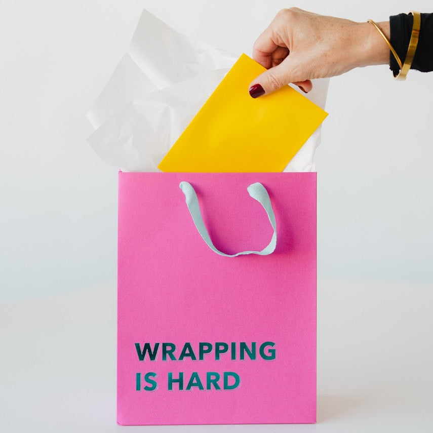 Pink gift bag with 'WRAPPING IS HARD' text, hand pulling yellow paper out.