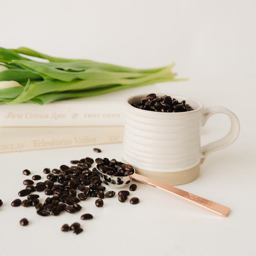 White mug with coffee beans, a spoon, and scattered coffee beans on a light surface.